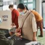 Voters casting ballots indoors on Election Day, wearing masks and using tablets for contactless voting.