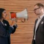 A woman using a megaphone to confront a man in a suit indoors, symbolizing political debate.