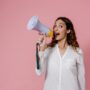 Woman in white shirt holding megaphone against a pink background, confidently speaking.