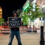 Man holds anti-cancer sign in a busy urban night setting, with light trails and buildings.